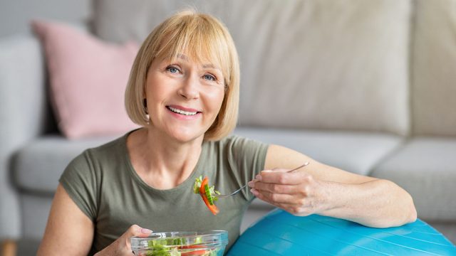 Exercise and healthy diet concept. Senior woman with fitball eating fresh vegetable salad at home. Mature Caucasian lady having veggies after sports training, keeping weight loss diet