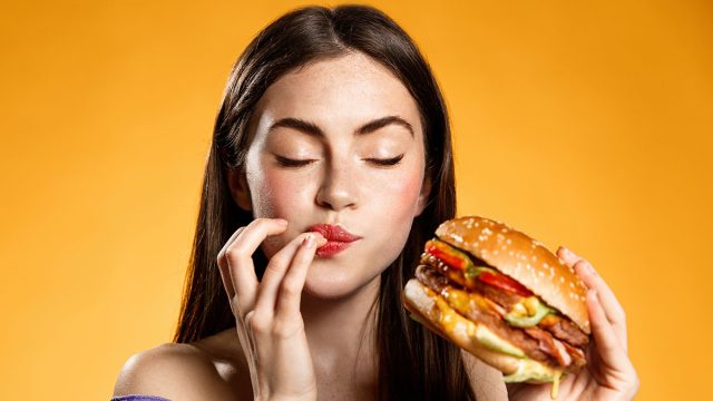 Woman eating cheeseburger with satisfaction. Girl enjoys tasty hamburger takeaway, licking fingers delicious bite of burger, order fastfood delivery while hungry, standing over orange background
