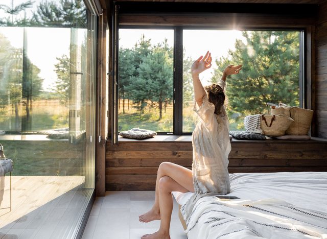 Woman wakes up in a country house or hotel with panoramic windows in pine forest raised her hands yawning. Good morning and recreation on nature concept