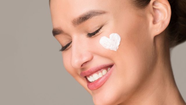A woman with a radiant smile is shown with a white cream applied on her face. She appears content and relaxed as she enjoys a skincare routine. The cream visibly hydrates and nourishes her skin.