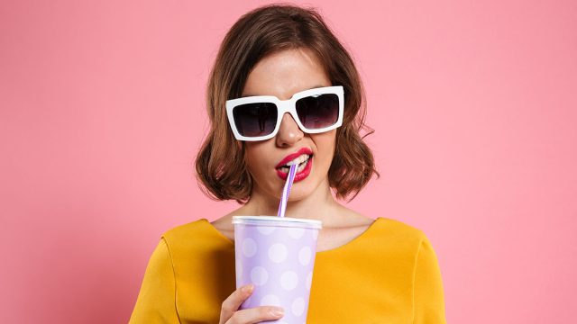 Portrait of a girl in sunglasses holding cup with drink and looking at camera isolated over pink background