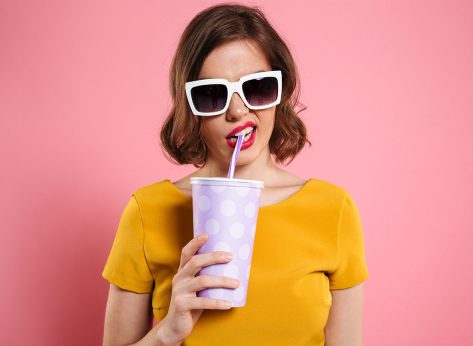 Portrait of a girl in sunglasses holding cup with drink and looking at camera isolated over pink background