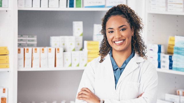 Pharmacy, pharmacist or portrait of woman with arms crossed or smile in customer services or clinic. Healthcare help desk, wellness or happy doctor smiling by medication on shelf in drugstore