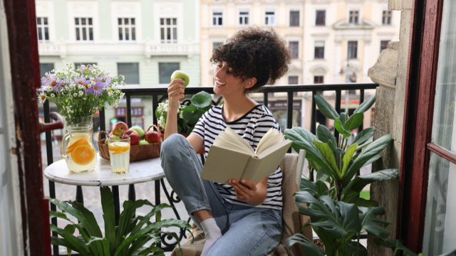 Young,Woman,Reading,Book,At,Table,On,Balcony,With,Beautiful