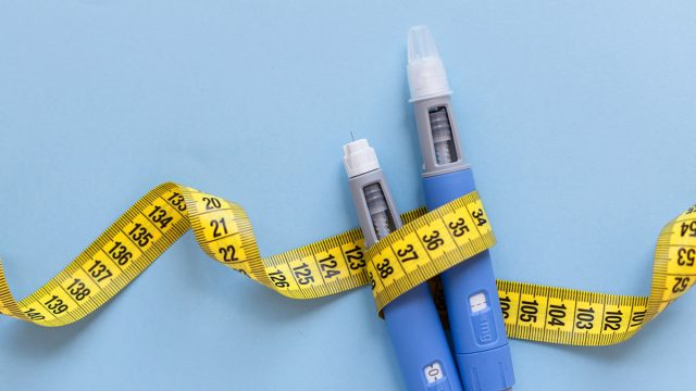 Two injectors dosing pens for subcutaneous injection of antidiabetic medication or anti-obesity medication hovering over a blue background. Yellow measuring tape around the injectors. antidiabetic