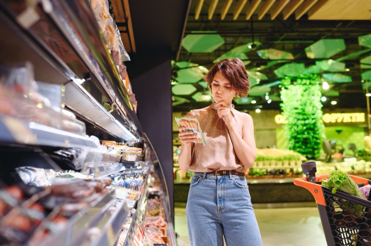 Young woman in casual clothes at supermarket store looking at meat sausage and read ingredients