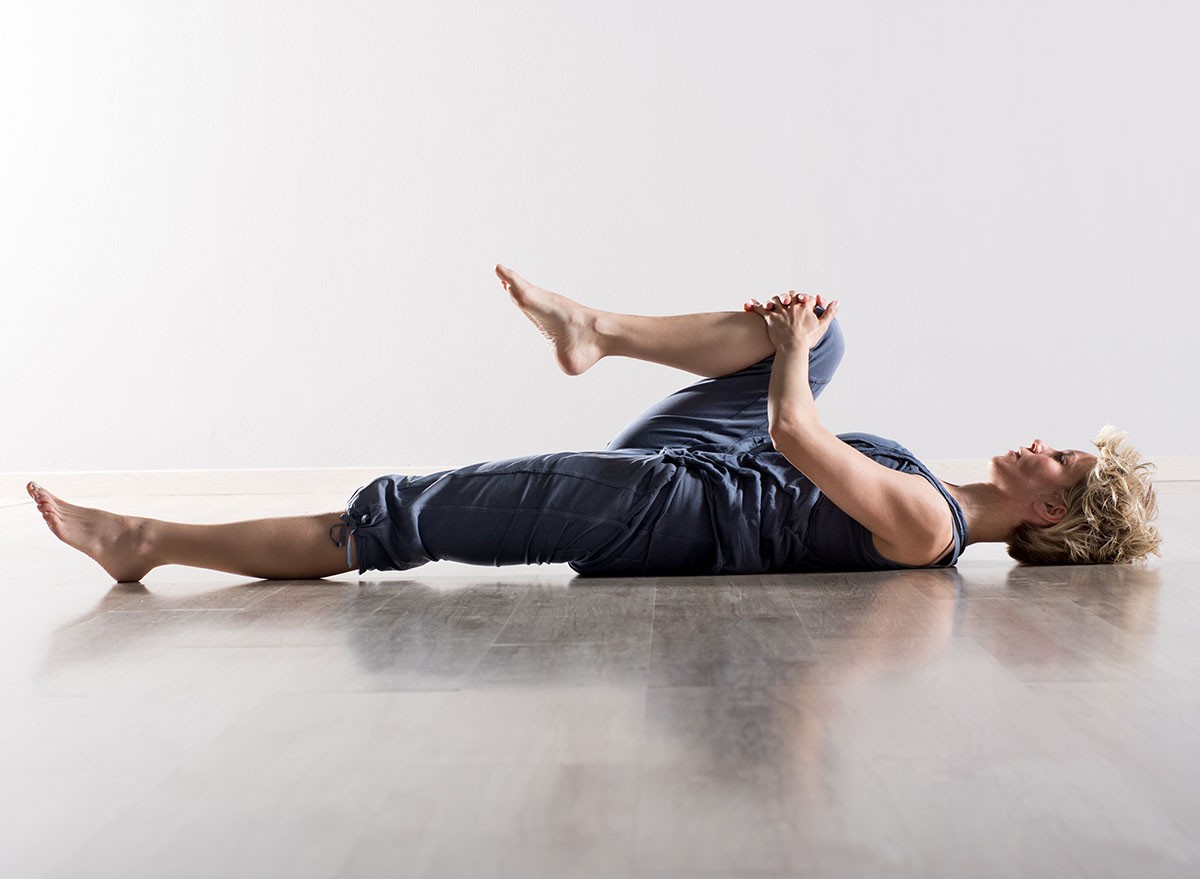 Single athletic woman with blond hair in blue outfit on back stretching hamstring muscles while holding knee close to her chest