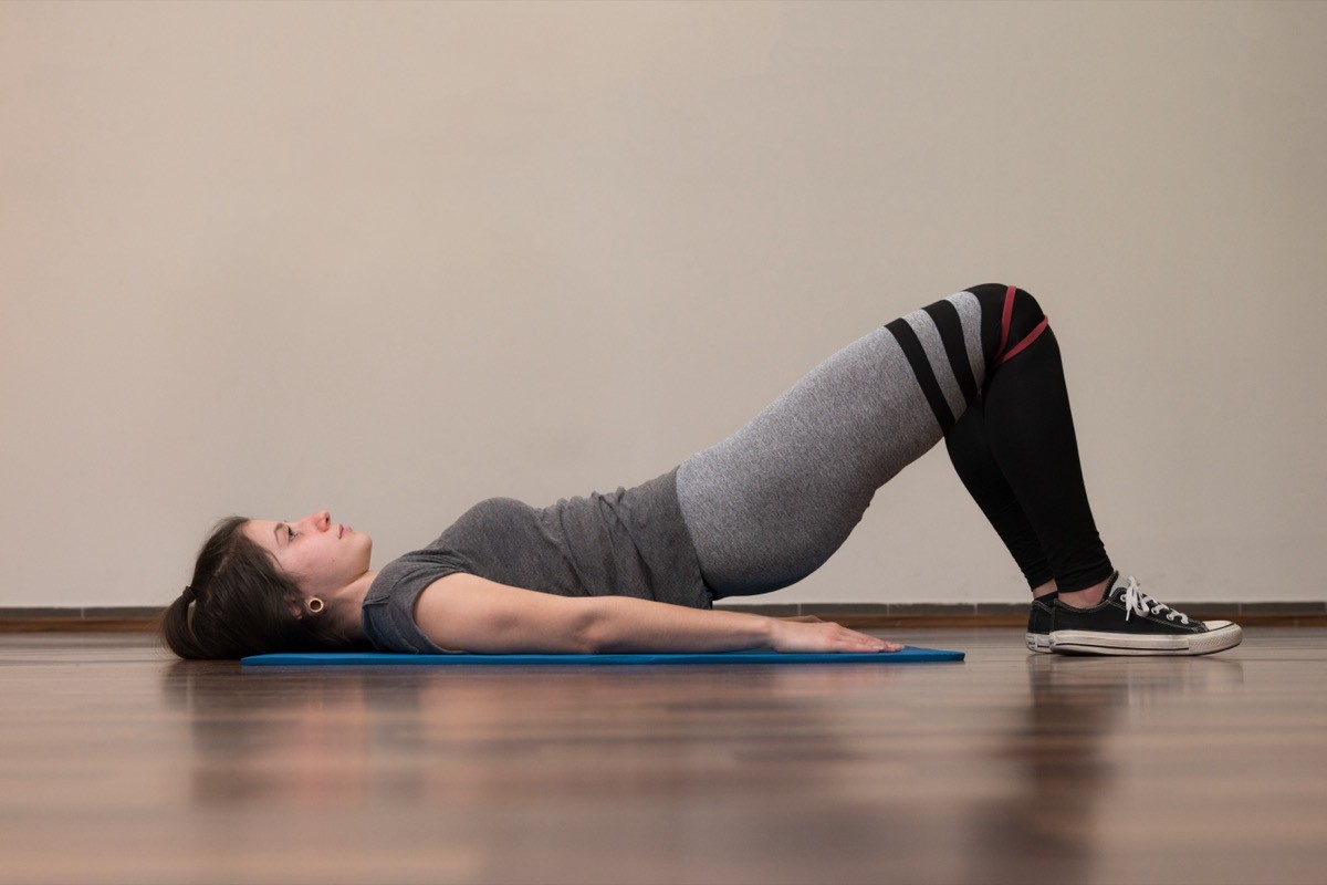 Young athlete girl performs glute bridge exercise with red rubber band in a gym.