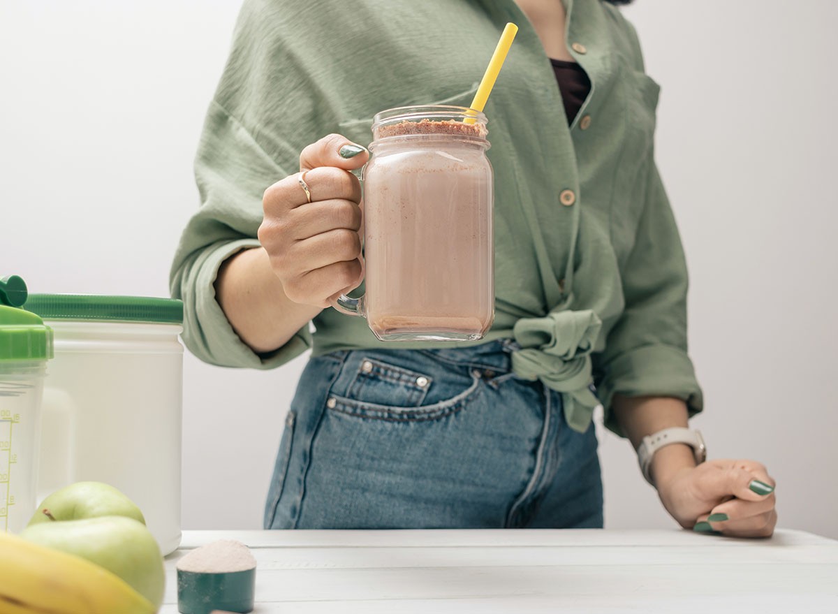 Young woman in jeans and shirt holding glass jar of protein drink cocktail, milkshake or smoothie above white wooden table with measuring spoon of protein powder, chocolate pieces, bananas and apples.