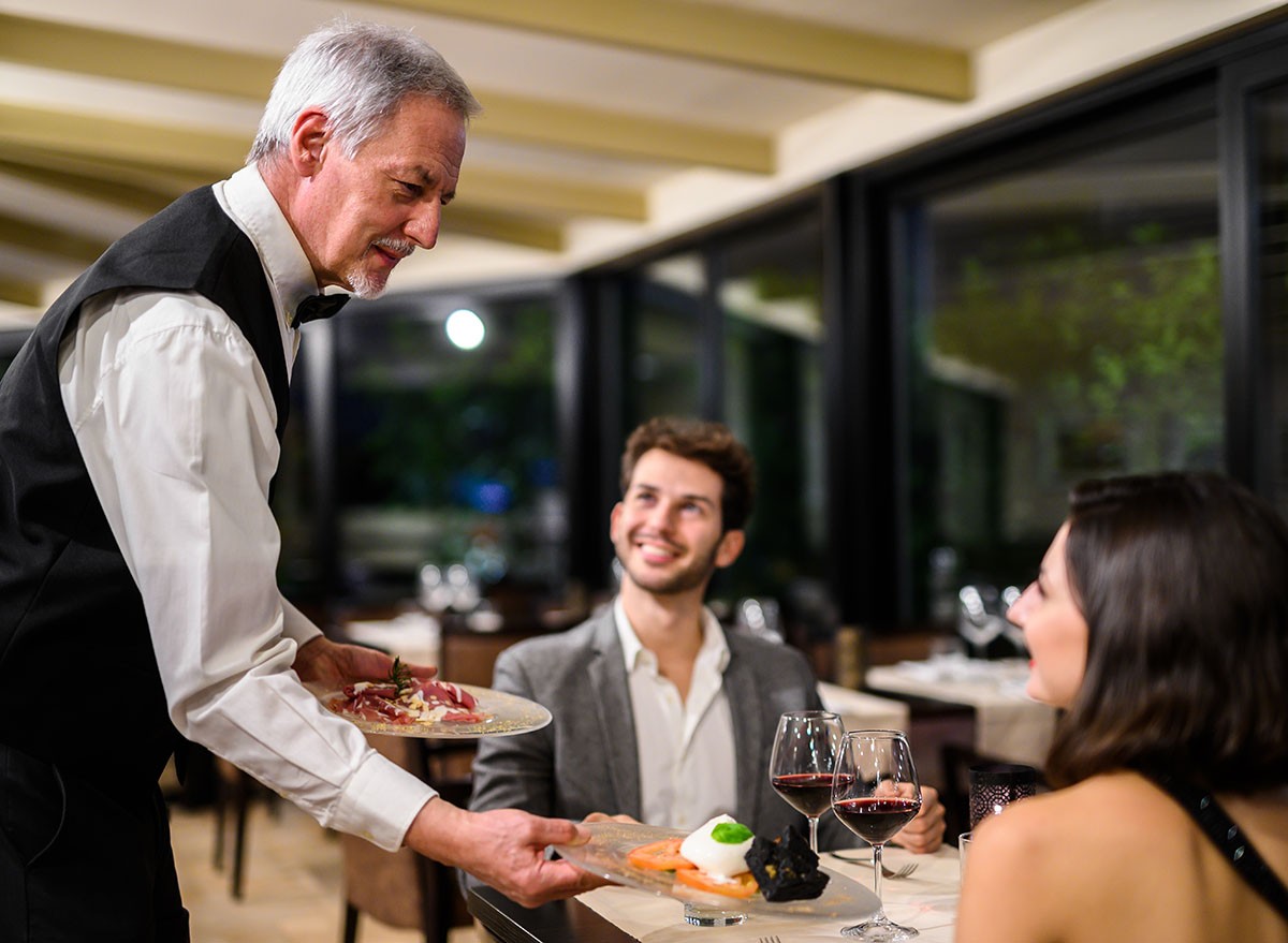 Professional senior male waiter presents a dish to a smiling young couple during a romantic dinner