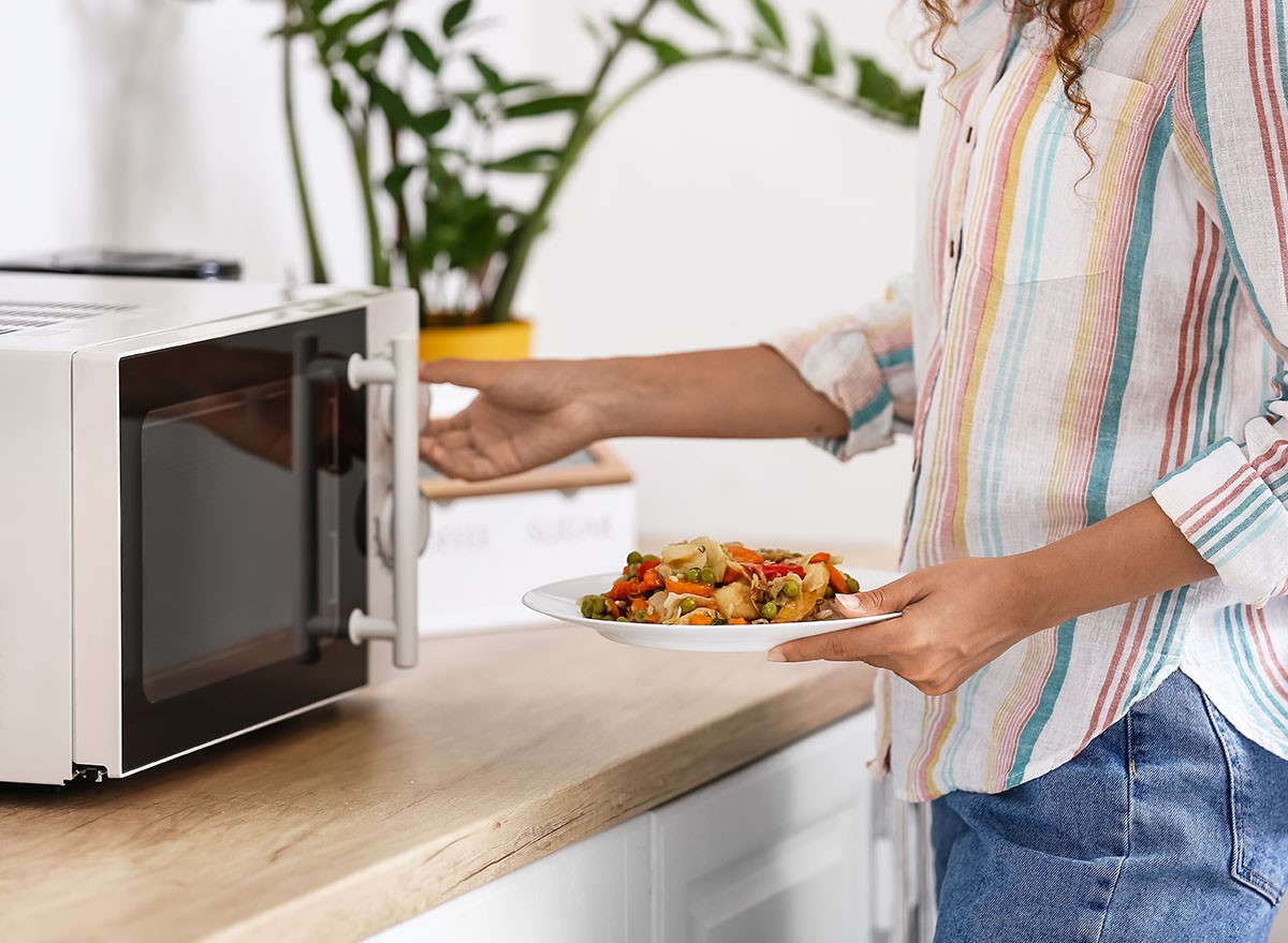 Young woman putting plate with food into microwave oven in kitchen