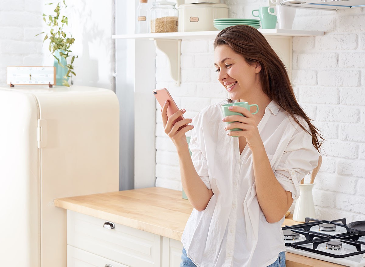 Young woman using smartphone leaning at kitchen table with coffee mug and organizer in a modern home. Smiling woman reading phone message. Brunette happy girl typing a text message