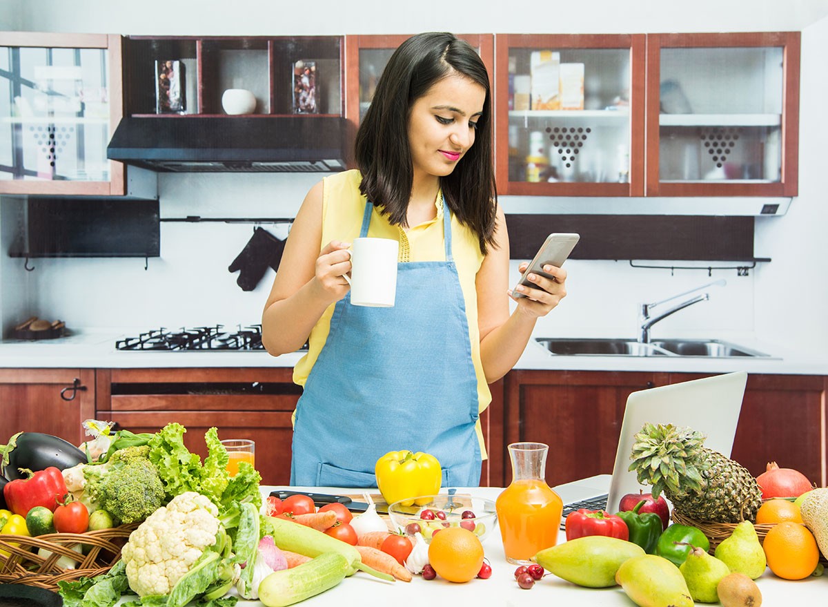 Attractive young Indian girl with apron in kitchen using mobile phone or smartphone with table full of fruits and vegetables and computer