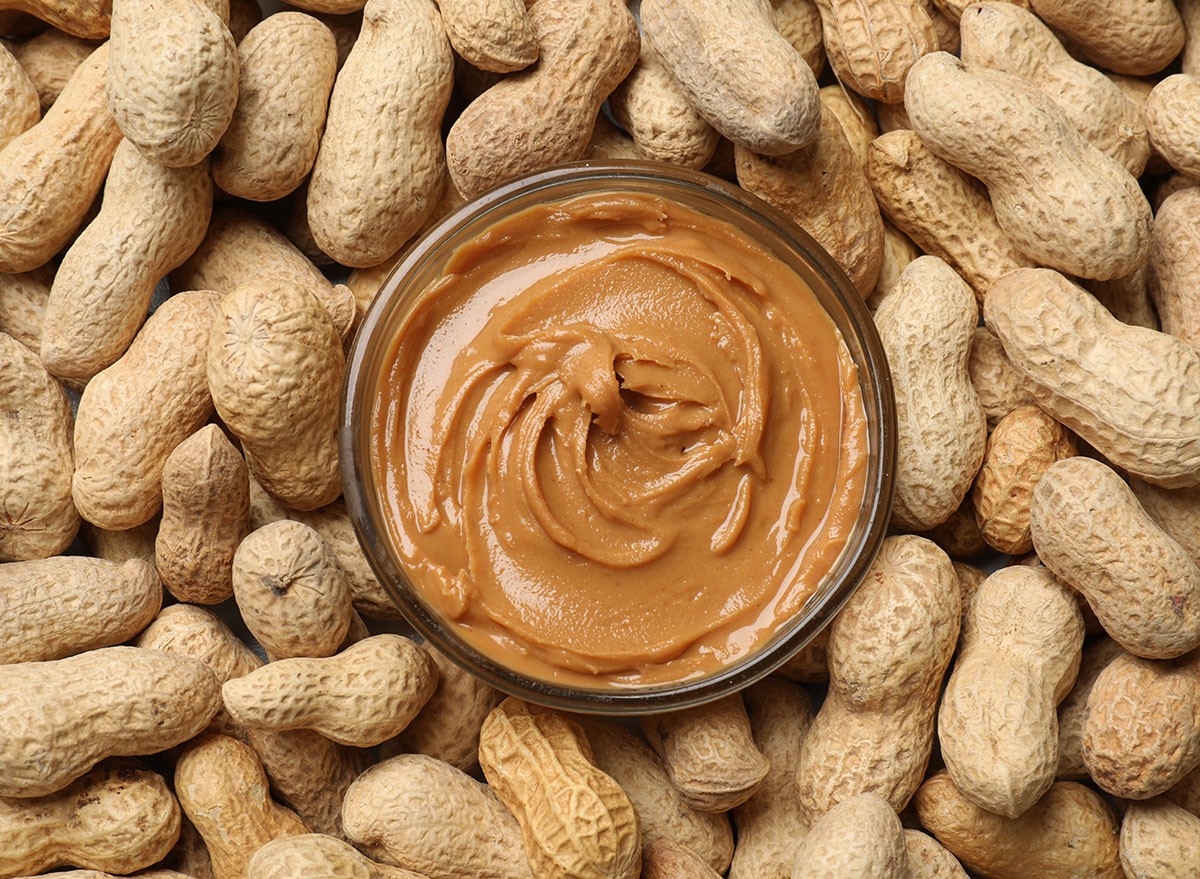 Tasty peanut butter in bowl among groundnuts, flat lay