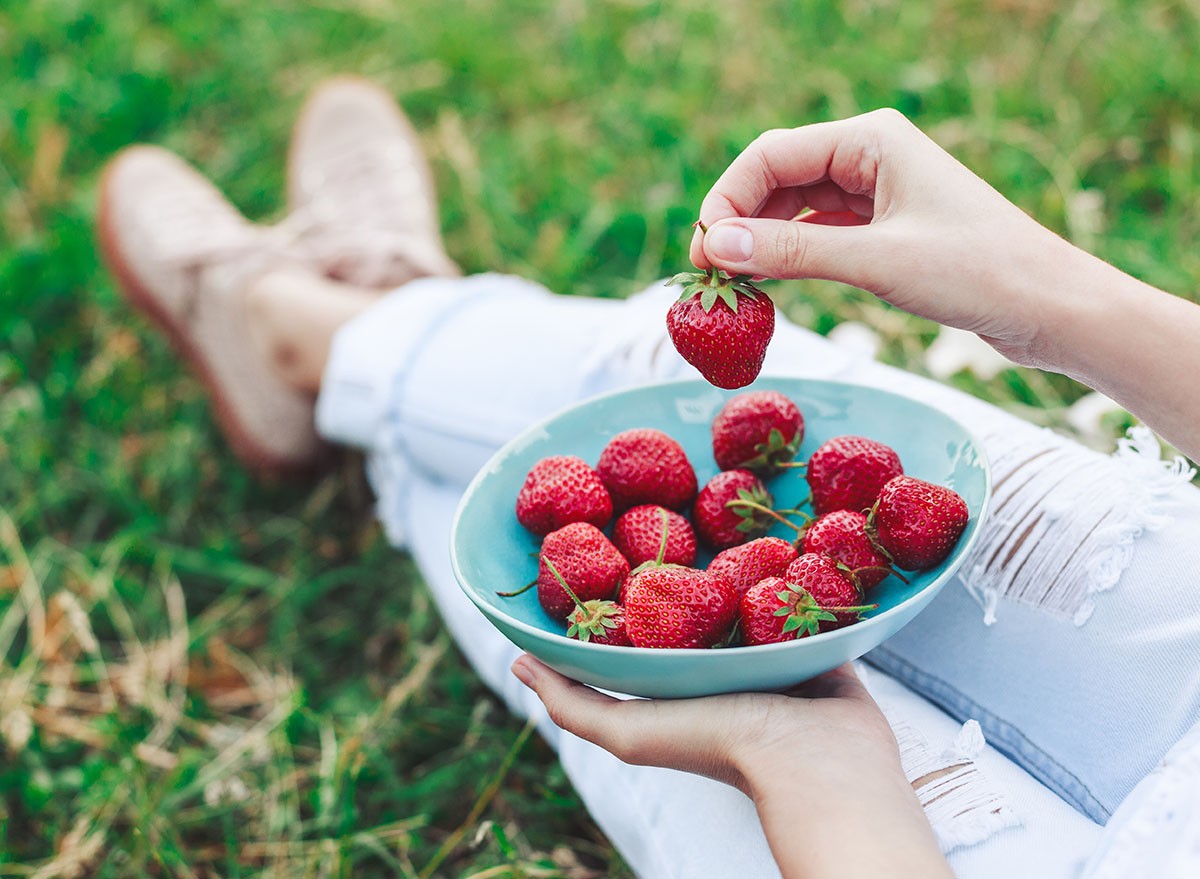 Girl in jeans sitting in summer grass and holding a plate of strawberries, knees and hands visible. Healthy breakfast, Clean eating, vegan food concept. Top view. Toning