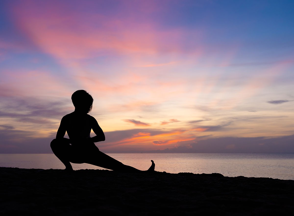 Silhouette of beautiful woman practicing yoga asana on the beach with sunrise on twilight blue vibrant sky and calm sea in background. Skandasana, Side lunge, Stretching, Power concept, Meditation.