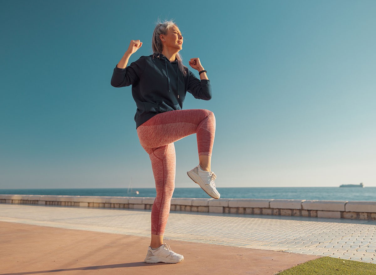 Woman in pink leggins and blue hoodie training, lifting one knee up and looking ahead on sunny day