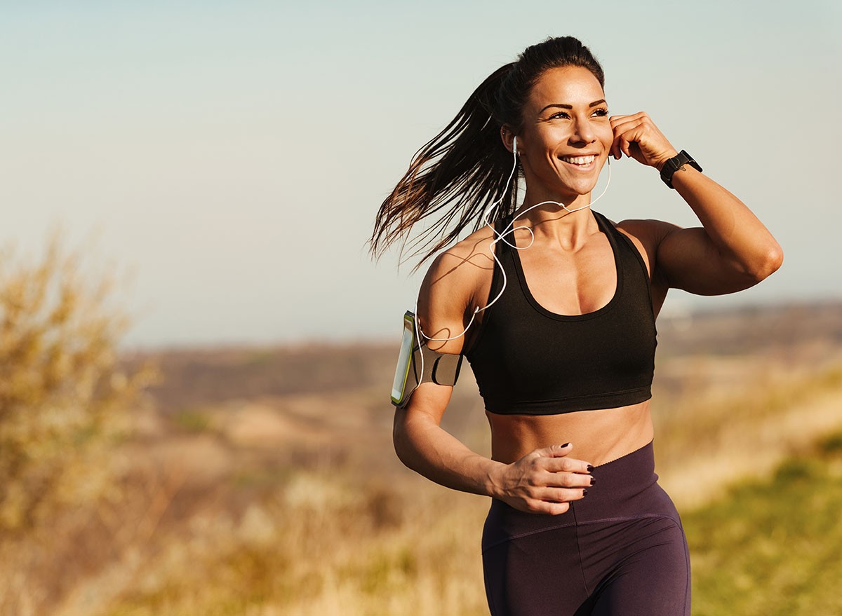 Happy muscular build woman listening music over earphones while running in nature. Copy space.