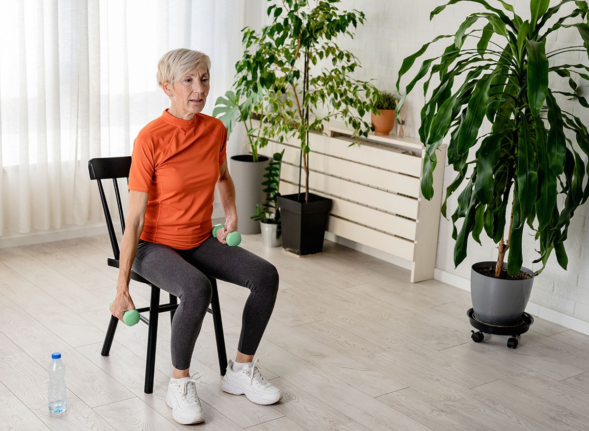 Smiling beautiful senior woman health instructor doing chair exercises with dumbbells