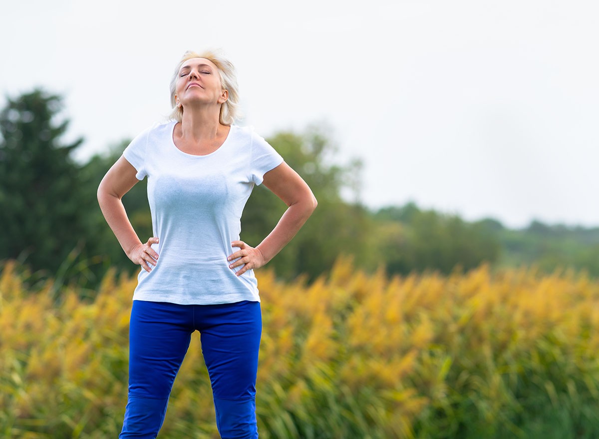 Adult woman with hands on hips looking upwards while wearing blue pants and white tee shirt with blurry yellow flowers in background