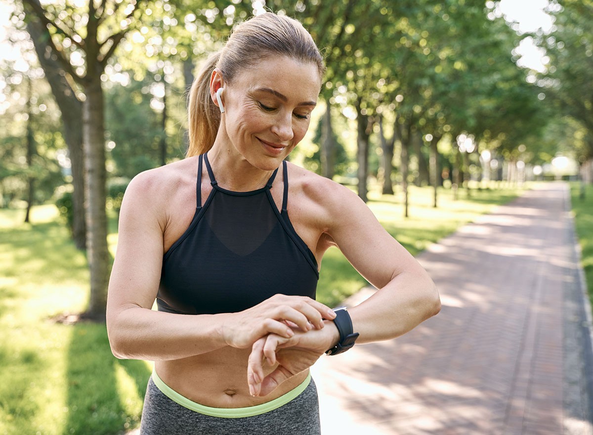 Morning jog. Beautiful sporty woman in sportswear and earphones looking at her watch, checking the result while running in a green park on a sunny summer day