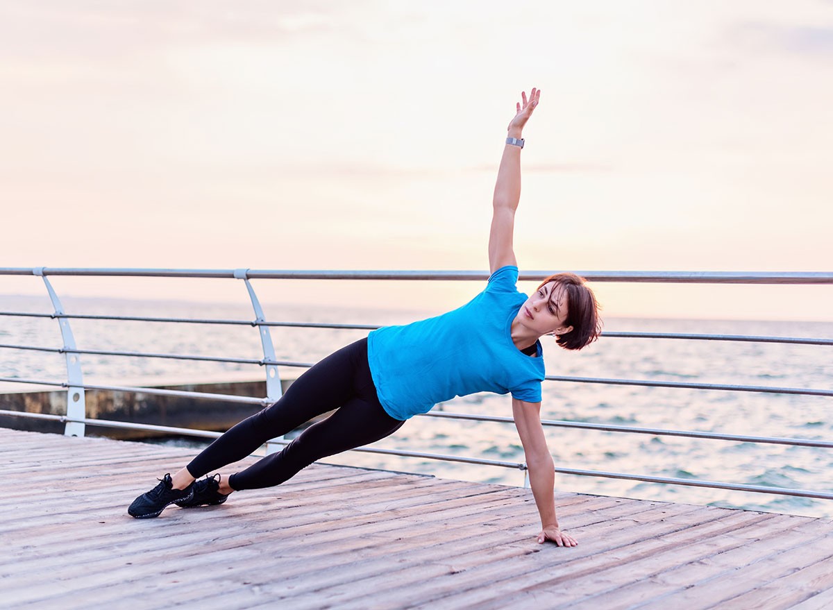 Attractive athletic woman in blue shirt doing arm plank rotation on seashore at sunrise