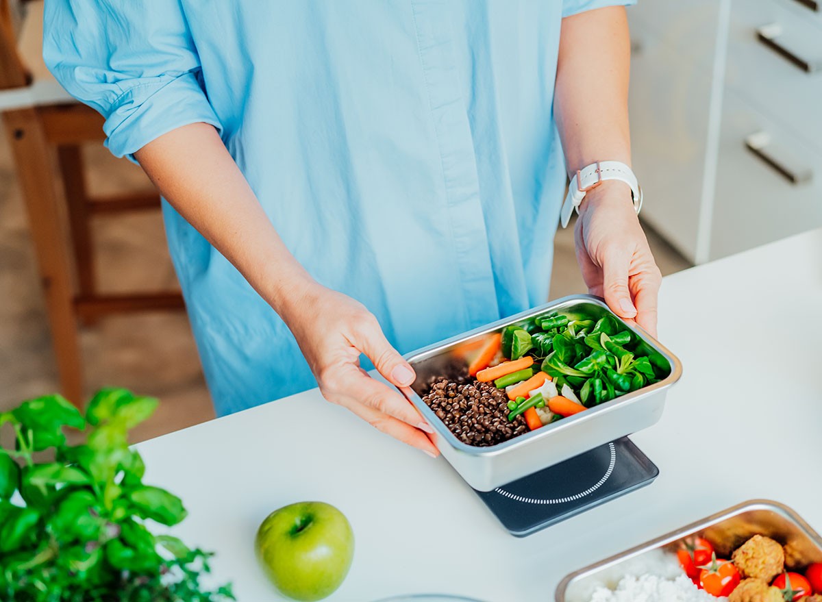 Healthy diet plan for weight loss, daily ready meal menu. Close up Woman weighing lunch box cooked in advance,ready to eat on kitchen scale. Balanced portion with healthy dish. Pre-cooking concept
