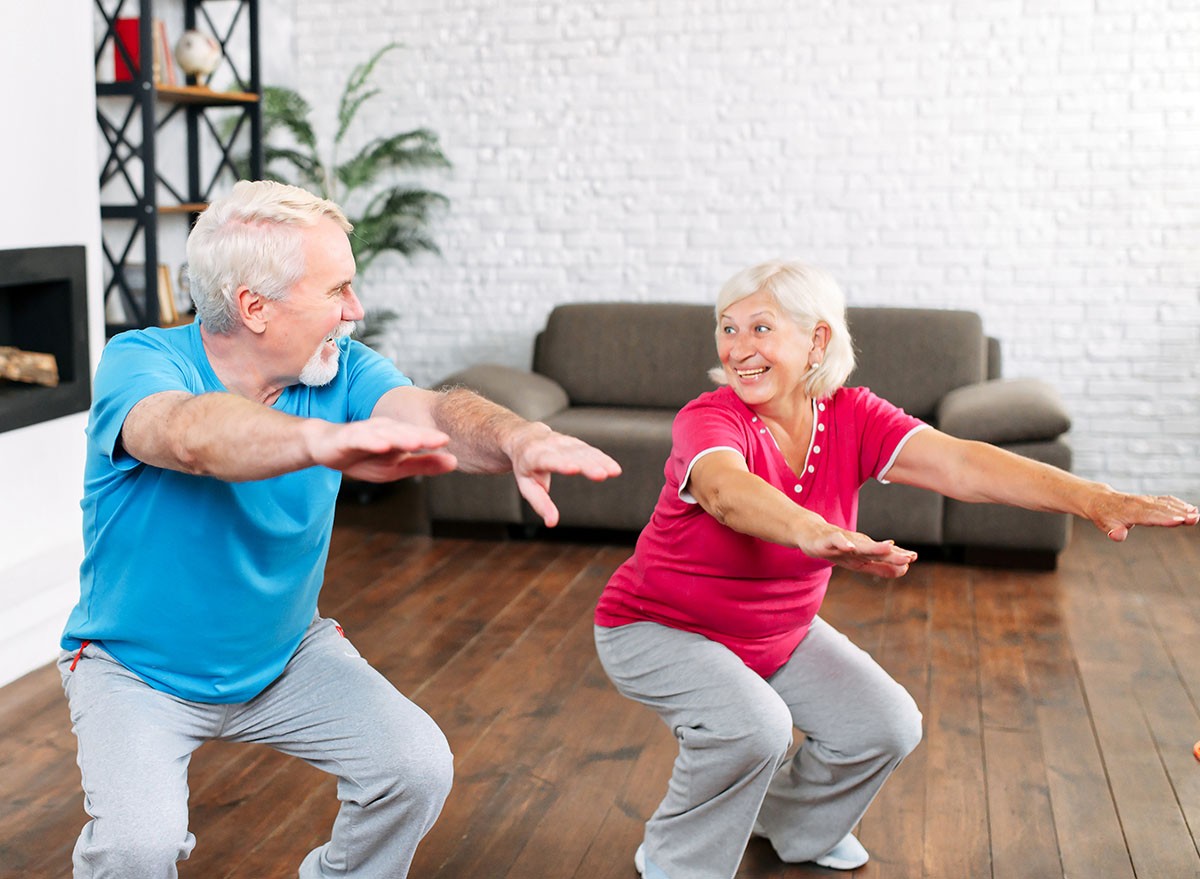 Elderly couple doing squats together at home. Cheerful grey-haired spouses doing exercises, look to each other and smiles, mature