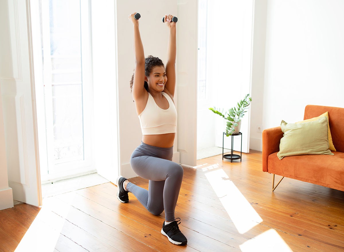 Cheerful athletic woman standing in lunge position and raising arms holding dumbbells, having bodybuilding workout in modern living room interior. Home fitness concept