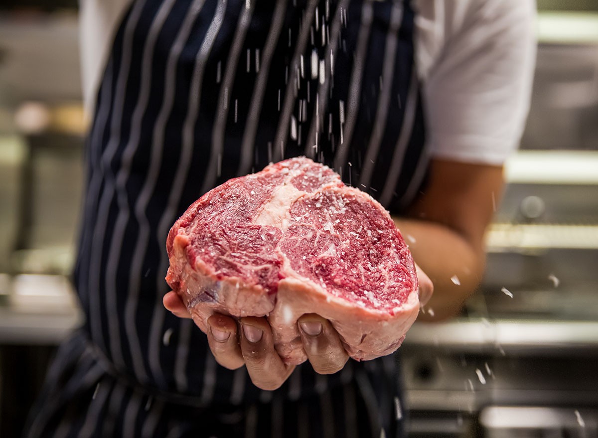 Close up of chef's hand seasoning a raw steak with salt crystals, beef, meat, protein, kitchen