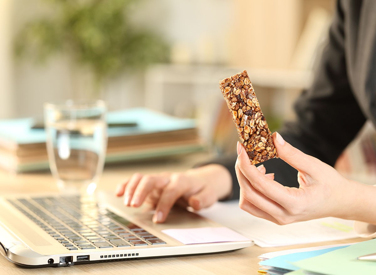 Close up of entrepreneur woman hands holding cereal snack bar working on laptop at home office
