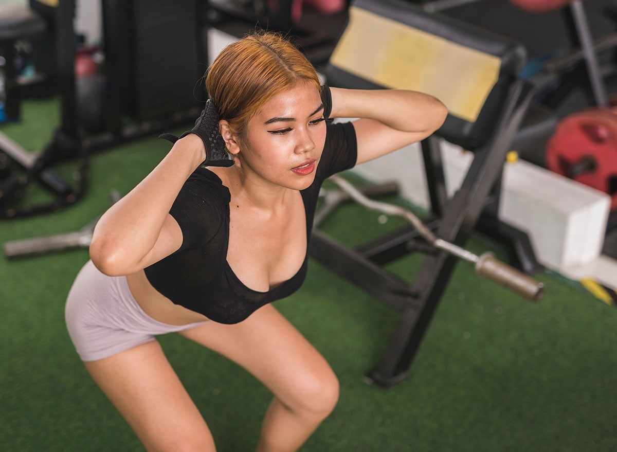 A young asian woman does prisoner squats at the gym. Hands on the back of the neck. Wearing a black crop top and fitness gloves. Training legs and lower body.
