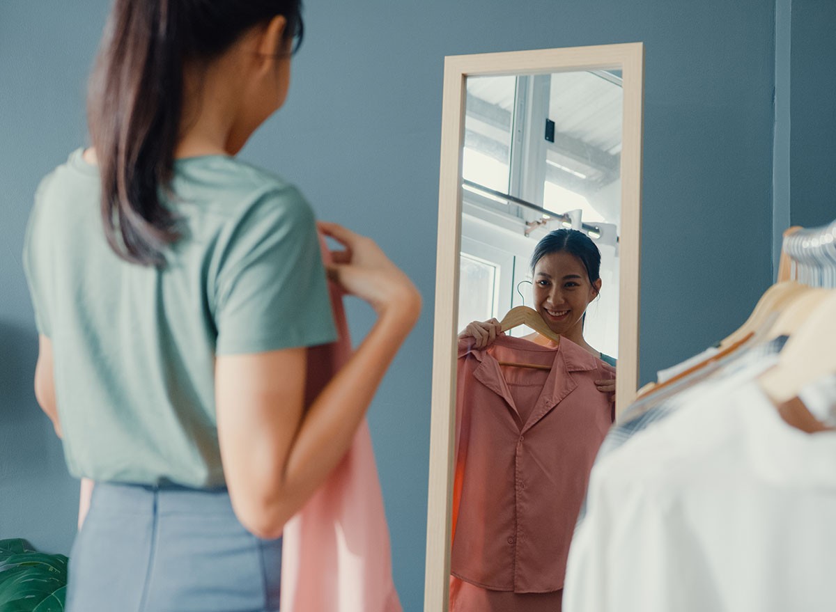 Beautiful attractive Asia lady choosing clothes on clothes rack dressing looking herself in mirror in living room at house. Girl think what to wear casual shirt. Lifestyle women relax at home concept.