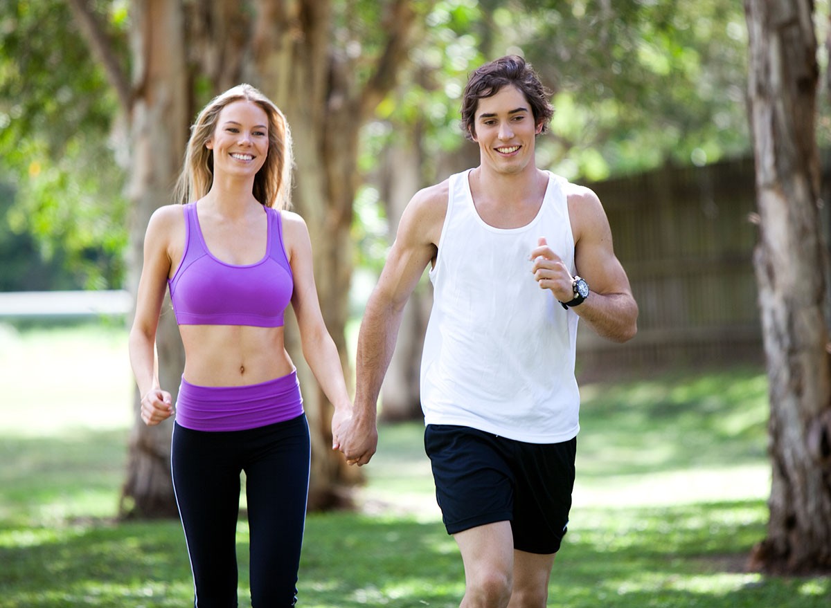 Portrait of a smiling young couple exercising in an outdoor setting while holding hands. The man is jogging, and the woman is walking. Horizontal shot. walk, walking